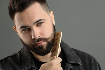 Handsome young man combing beard on grey background