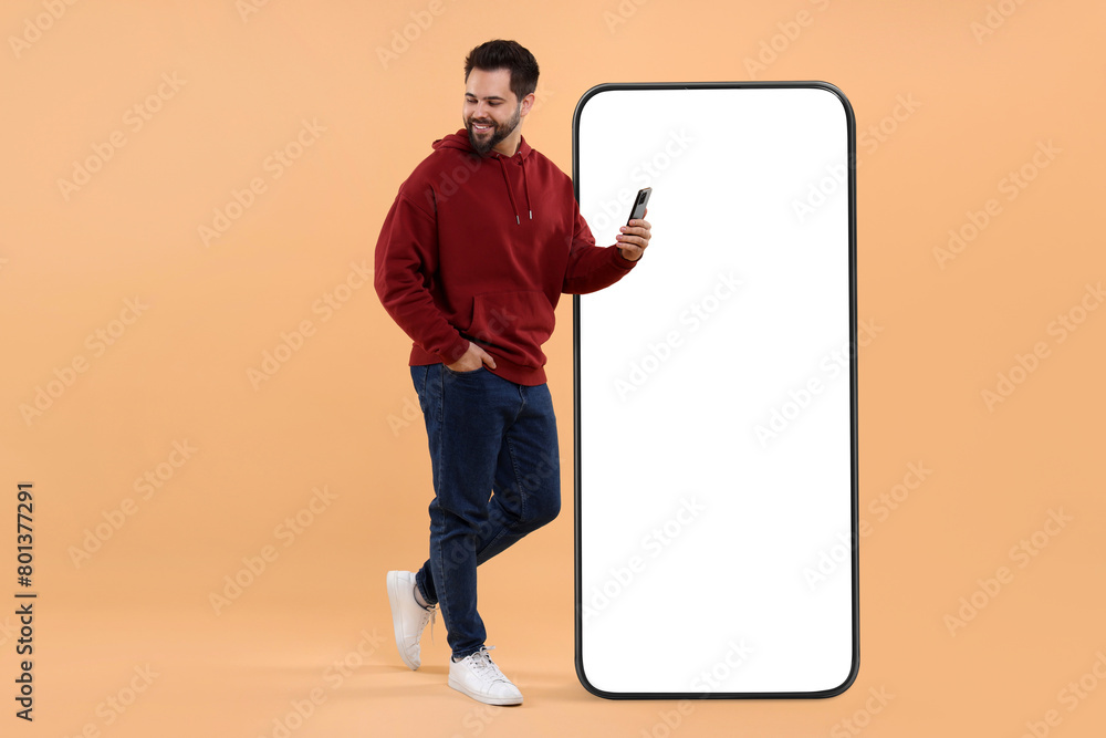 Aufkleber man with mobile phone standing near huge device with empty screen on dark beige background. mockup f