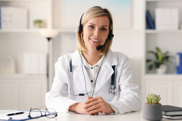 Portrait of smiling doctor in headphones having online consultation at table indoors