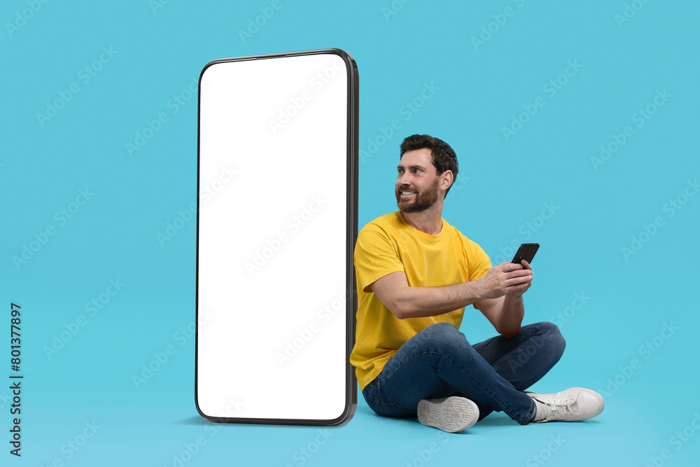 Poster man with mobile phone sitting near huge device with empty screen on light blue background. mockup fo