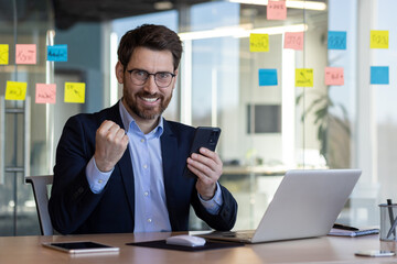 Professional male executive in a suit happily celebrates a business achievement using his smartphone at a modern office desk with post-it notes.