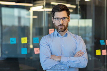 A professional male executive stands confidently with arms crossed in a contemporary office space, surrounded by glass walls and sticky notes.
