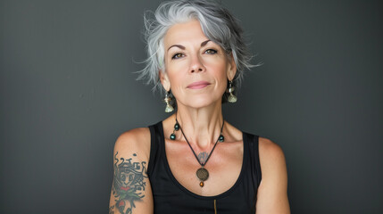 Naklejka premium Captivating photo of an attractive middle-aged woman with grey hair and tattoos, wearing earrings, necklace, and a black tank top.