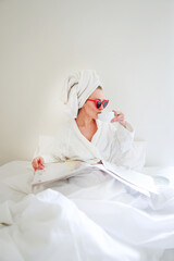 Morning. A girl in a white robe and a towel on her head put on glasses in the shape of a heart and reads a newspaper. Copy space