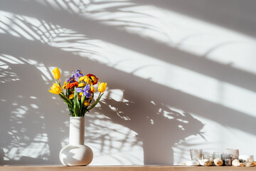 Scandinavian home interior with colorful bouquet of various flowers in ceramic vase standing on wooden cabinet under sunlight and shadows on white gray wall. Minimalist design of home decor.