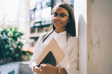 Portrait of cheerful african american young woman dressed in trendy white shirt smiling at camera while holding folder and modern smartphone in hands standing outdoors near office building