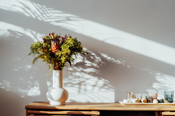 Scandinavian home interior with with exotic protea flowers bouquet in ceramic vase standing on wooden cabinet under sunlight and shadows on white gray wall. Minimalist design of home decor.
