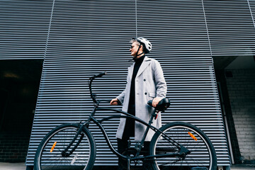 Stylish male in coat, sunglasses and protective helmet standing with retro bicycle near gray urban...