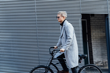 Stylish male in checked coat and sunglasses riding on retro bicycle near gray urban wall in sunny day. Neutral carbon footprint transportation. Green eco friendly mobility sustainable transport - Powered by Adobe