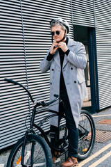 Stylish male in coat, sunglasses putting protective helmet on head before going to go ride by bicycle in city street. Neutral carbon footprint. Green eco friendly mobility sustainable transport.