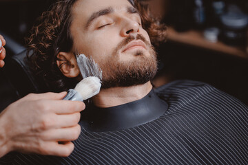 Barber applies shaving foam with brush to man beard in barbershop, warm toning