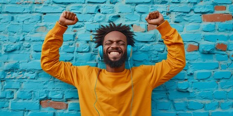 Joyful man with headphones dances to the rhythm, raising his fists in victory against a contrasting blue brick wall background