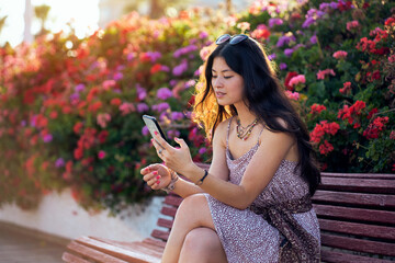 A woman is sitting on a bench in a park using her mobile phone. She is smiling and looking at the screen scrolling through social networks.