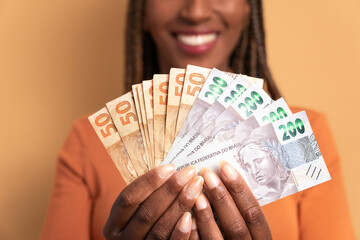 anonymous afro brazilian woman with money, brazilian real in beige studio background. finance,...