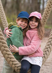  girl tenderly hugs her brother. Children sit on a swing in a city park and laugh. Brother and...