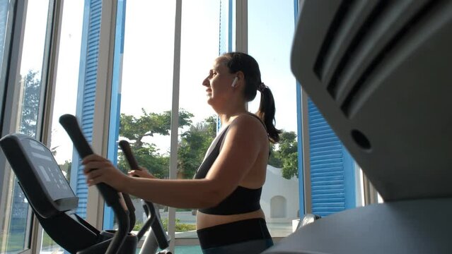Young woman listening to music in headphones and working out on elliptical trainer at gym, slow motion. Healthy lifestyle concept