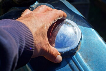 hand unscrews the round chrome shiny cap from a blue iron old dirty motorcycle tank during the day...