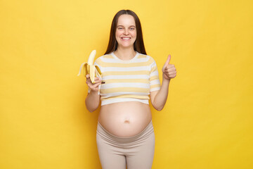 Joyful Caucasian pregnant woman with bare belly wearing casual top isolated over yellow background...