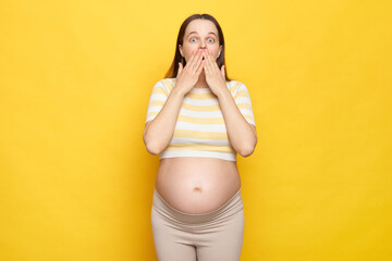 Shocked astonished Caucasian pregnant woman with bare belly wearing casual top isolated over yellow...