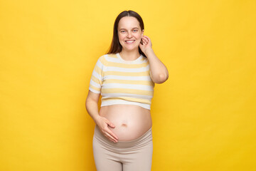 Beautiful happy delighted Caucasian pregnant woman with bare belly wearing casual top isolated over...