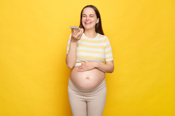 Cheerful joyful Caucasian pregnant woman with bare belly wearing casual top holding mobile phone...