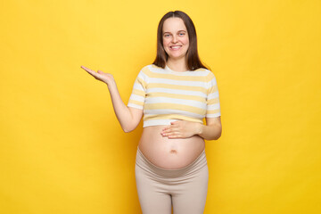 Smiling happy Caucasian pregnant woman with bare belly wearing casual top isolated over yellow...