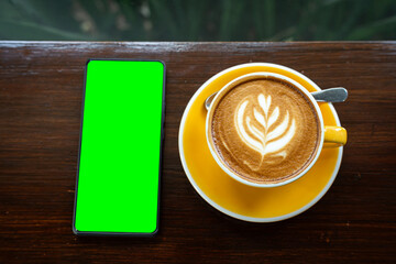 Hot coffee latte with latte art milk foam in cup mug,smartphone with empty screen on wood desk on...