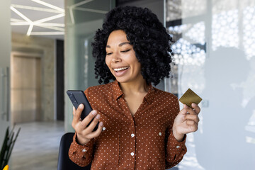 A joyful African American woman uses her smartphone and credit card to shop online. She stands in a...