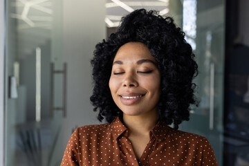 A young African-American woman with curly hair, dressed in a polka dot shirt, closes her eyes and smiles, enjoying a serene moment in a sunlit modern office environment.