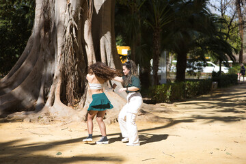 Latin women couple, young, dancing bachata with a big tree in the background in an outdoor park,...