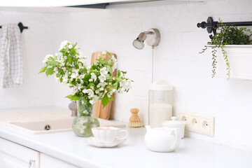 Spring morning in light kitchen: bouquet of white flowers, white teapot and glass of green tea.