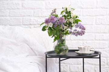 Spring morning in cosy bedroom: coffee table with bouquet of different flowers, cup of coffee, white bed in light room.