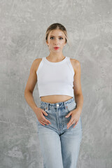 Caucasian woman is wearing a white top and blue jeans. She stands against a grey background in the studio