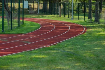 a curve of a red running track of an athletics stadium with three lanes between green grass