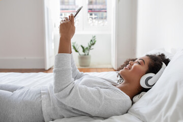 Hispanic woman is laying comfortably on a bed, wearing headphones and listening to music. She...