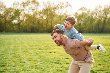Sitting on man's shoulders. Happy father with son are having fun on the field at summertime