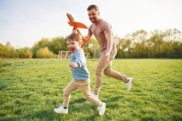 Toy plane in hand, running. Happy father with son are having fun on the field at summertime