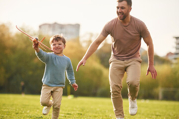 In casual clothes. Running with toy plane. Happy father with son are having fun on the field at...