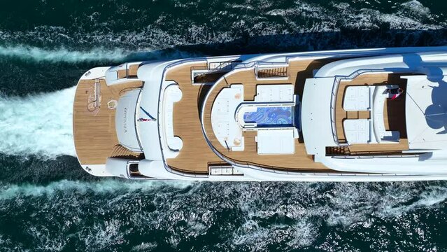 Aerial drone top down tracking video of beautiful modern super yacht with wooden deck and helipad cruising in high speed deep blue open ocean sea