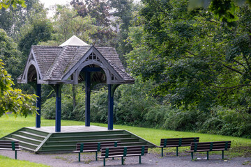 A wooden open-air amphitheater in a park. There are steps leading up to the platform with a...