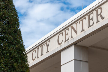 A city center sign on the exterior of a cream colored building with an overhang and square column. The recreation facility has a large green hedge in front with a blue sky in the background.