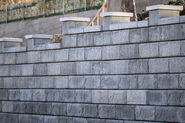 A large grey precast concrete block wall. The massive rectangle shaped bricks are interlocking cinder bricks with granular backfill. The textured castle style wall has large square caps. 