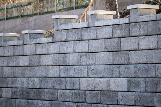 A large grey precast concrete block wall. The massive rectangle shaped bricks are interlocking cinder bricks with granular backfill. The textured castle style wall has large square caps. 