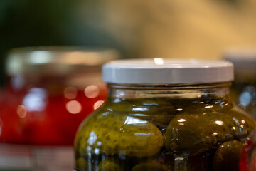 A glass jar filled with green pickled cucumber with a white metal lid. A second glass crock of red peppers with a gold metal lid.The food has pickling spices, vinegar, and garlic  in the canned bottle