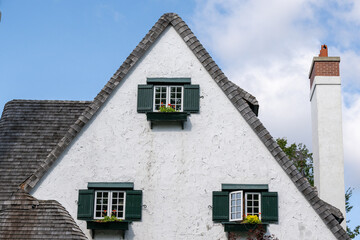 The exterior peaked wall of a large white stucco vintage house with grey weathered and worn cedar roof shakes. There's a small box filled with flowers under each glass window with green shutters.