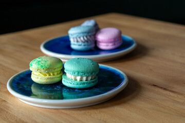 Five French macarons; pink, green, blue, and yellow color. The assorted elegant sandwich style sweet desserts lie on blue and brown ceramic plates and a wooden table. The meringue pastries are round.