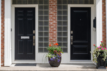 Two black metal doors with flower pots on the steps of duplex houses. It has a red brick wall with white wooden trim. There are glass blocks for windows on both sides of the building entrances.