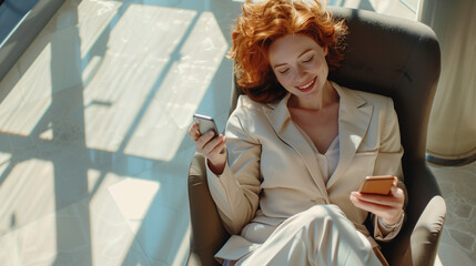 Smiling young professional business woman with red hair holding mobile cell phone in hands relaxing at work sitting in chair in sunny office looking away using smartphone, top view from above