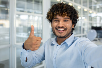 Confident young man with curly hair showing a thumbs up while taking a selfie at a bright, well-lit office setting. Positive and professional vibe.