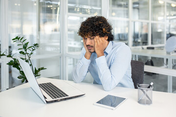 Young businessman with curly hair feeling stressed and tired while working at his laptop in a...